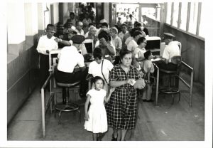 Black and white photograph; A women looks at the camera and so does a young girl to her right. There is a line of people behinf them.