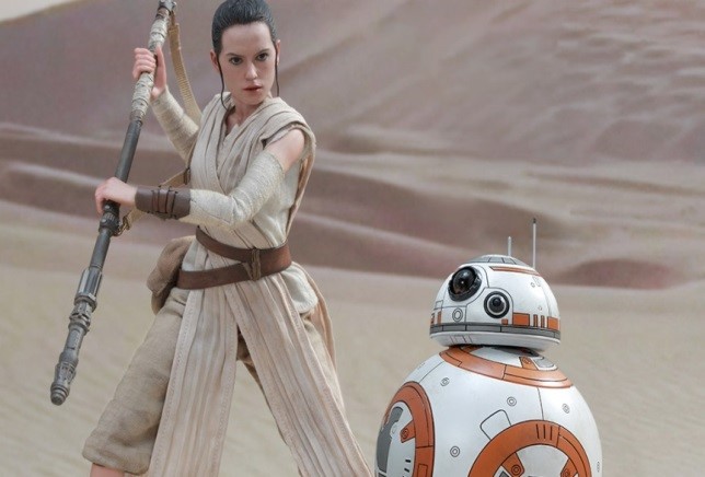 Rey stands in a desert looking down at a white and orange robot.