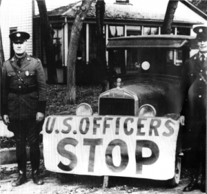 A black and white photograph of two police officers standing in front of a truck with a white sign on it that reads "U.S. Officers Stop"