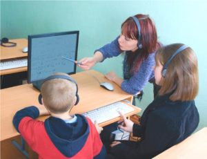 Children sit at a desk with headphones on while looking at a computer. A teacher with headphones on points to the computer.