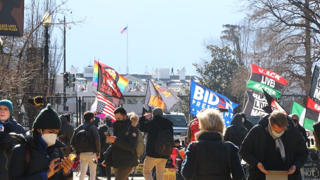 Daytime street-level view of protesters in front of the fence in front of the White House. Several are flying flags, including Black Lives Matter, Biden for president, and the black power fist set against the gay pride flag.